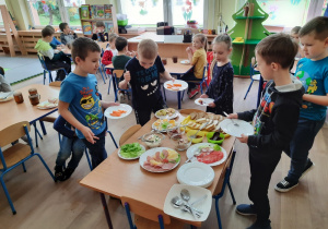 Pięcioro dzieci dokonuje wyboru potraw ze stołu szewdzkiego, w tle dzieci siedzą przy stolikach i spożywają śniadanie